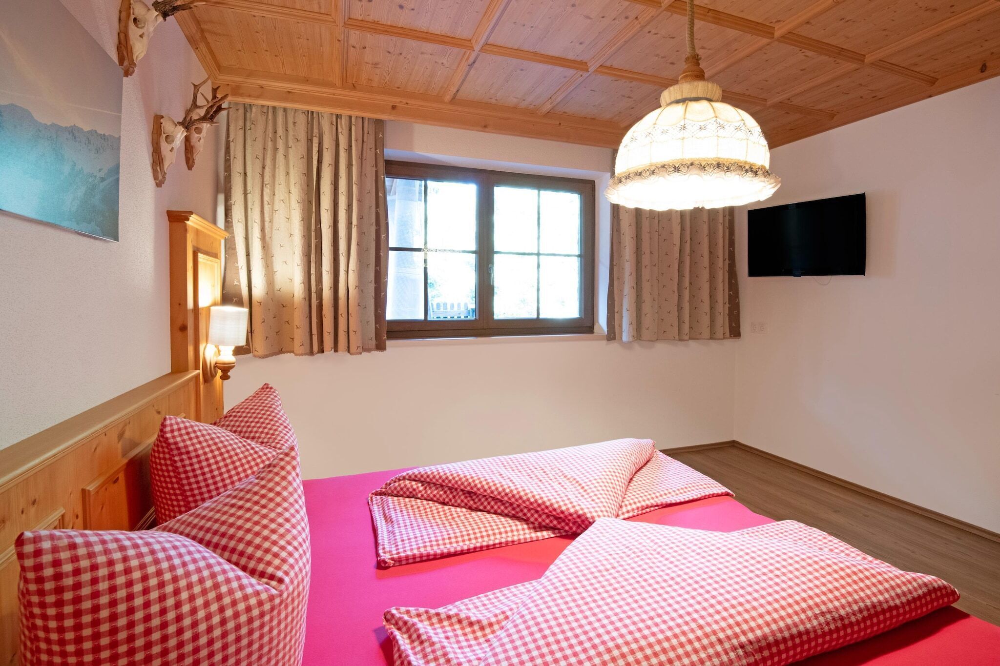 Une pièce avec un plafond en bois, un lit recouvert d’une couette à carreaux rouges, une fenêtre avec des rideaux, un téléviseur fixé au mur et une lampe fixée au mur.