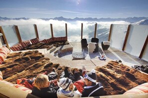 Un salon situé en altitude, avec des sièges disposés en cercle autour d’un foyer central, et offrant une vue panoramique sur les montagnes.