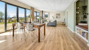 A spacious living area with a wooden dining table, chairs, and a sofa. There is a large window providing a view of the outdoors. - "Edvia" - 200m to the fjord by Interhome (Holbæk)