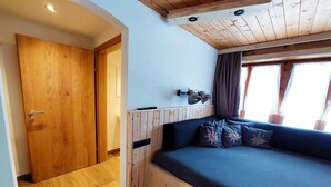 Una habitación con techo de madera, cama con ropa oscura, lámpara de pared y ventana con cortinas.