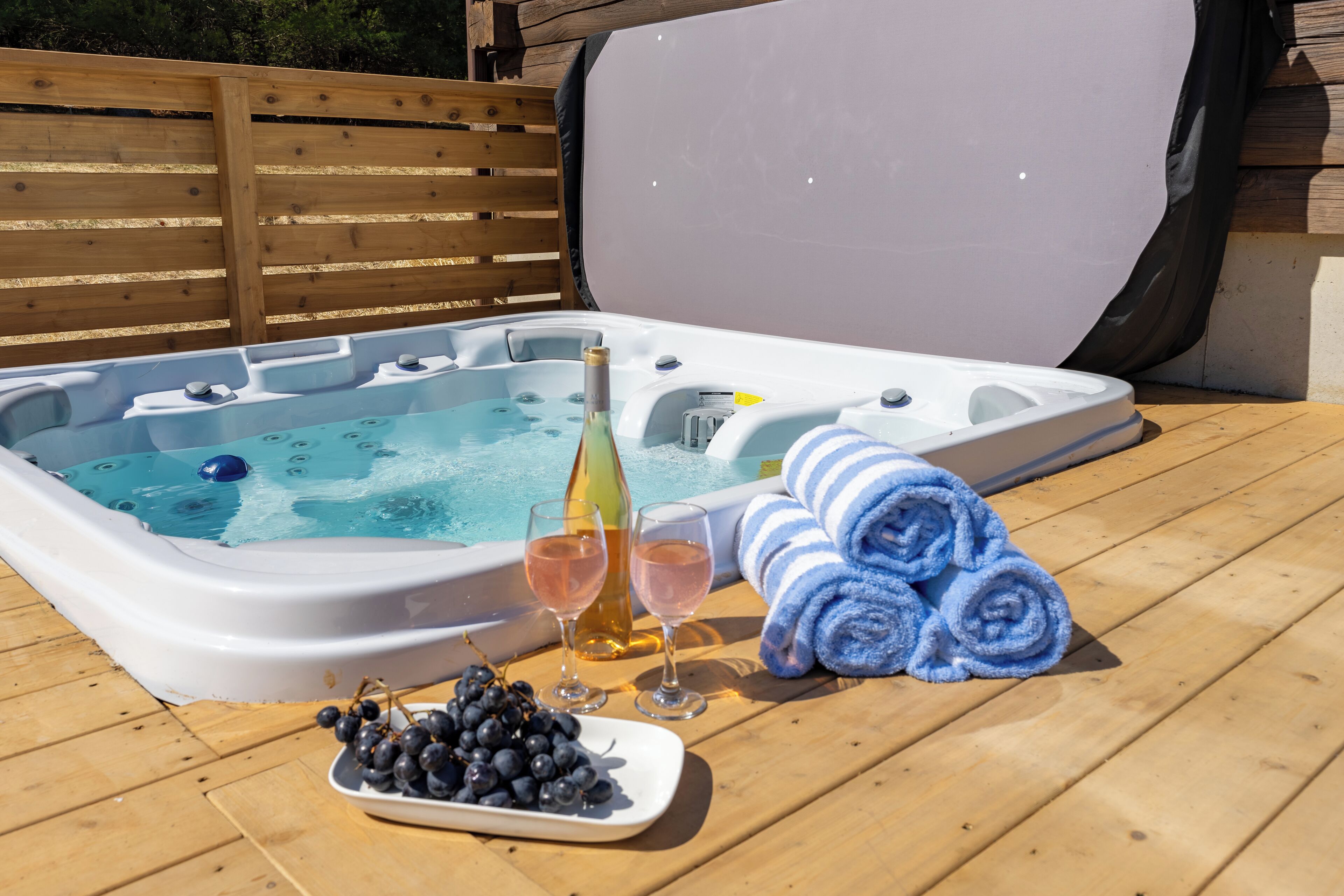 A hot tub with a wooden deck, two glasses of wine, a bottle of champagne, and a plate of grapes.