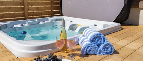 A hot tub with a wooden deck, two glasses of wine, a bottle of champagne, and a plate of grapes.