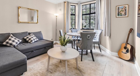 A living room with a grey sofa, a round white table, and a dining area with chairs. - Stylish 3-Bedroom Flat in Belsize Park – Perfect for Families & Business Trips! (Belsize Park)