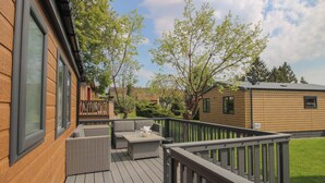 Una terraza de madera con mobiliario exterior, barandilla y vista a una zona residencial con árboles y casas.