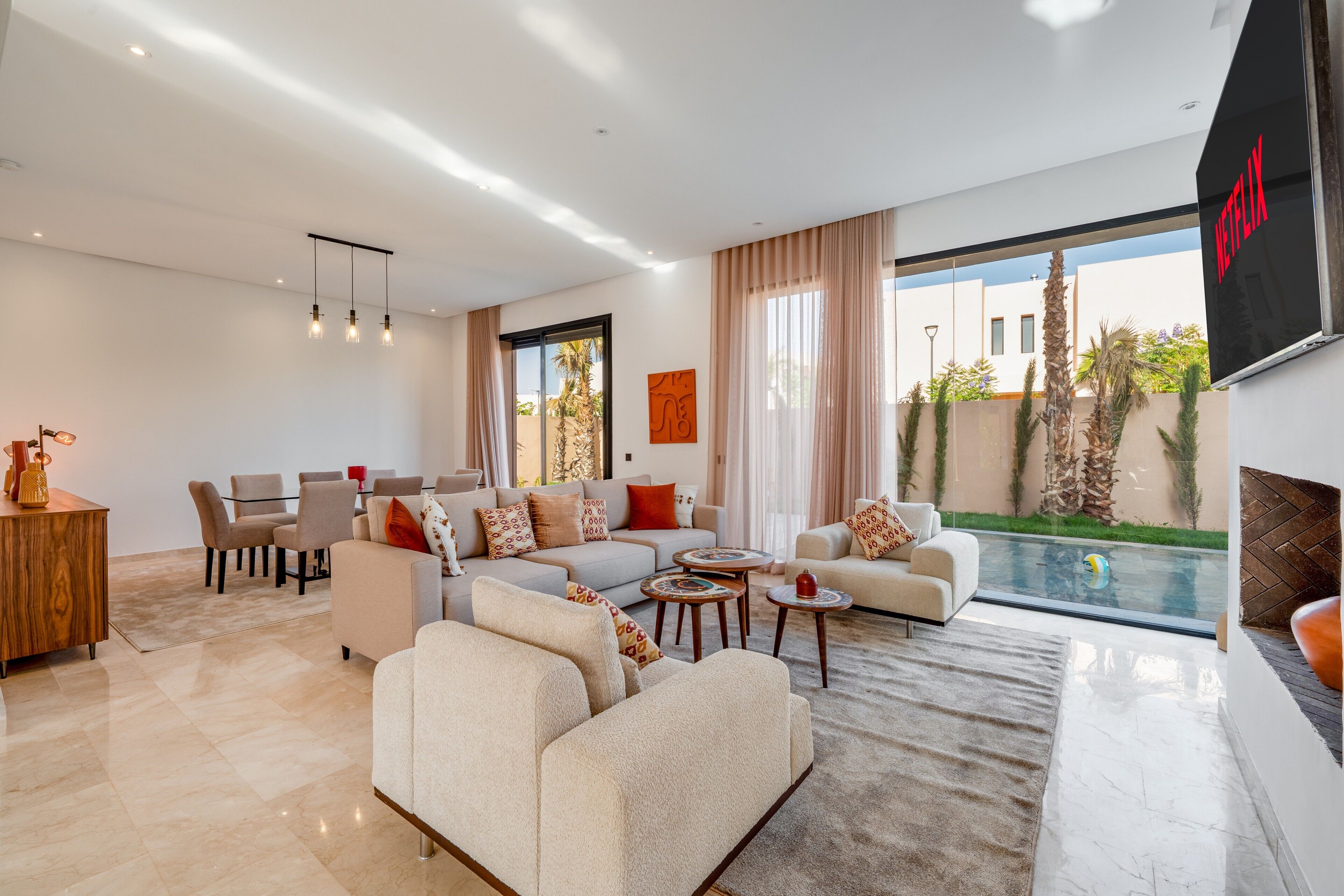 A modern living room with a pool view, a dining area, and a TV screen displaying the Netflix logo.