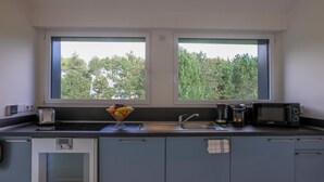 Una cocina compacta con microondas, tostadora y horno, un bol de fruta en la encimera, y vistas a los árboles a través de las ventanas.