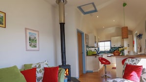 Una sala de estar acogedora con un sofá verde, cojines rojos y una alfombra roja. Una estufa de leña negra se encuentra en una esquina, y se ve una cocina al fondo.