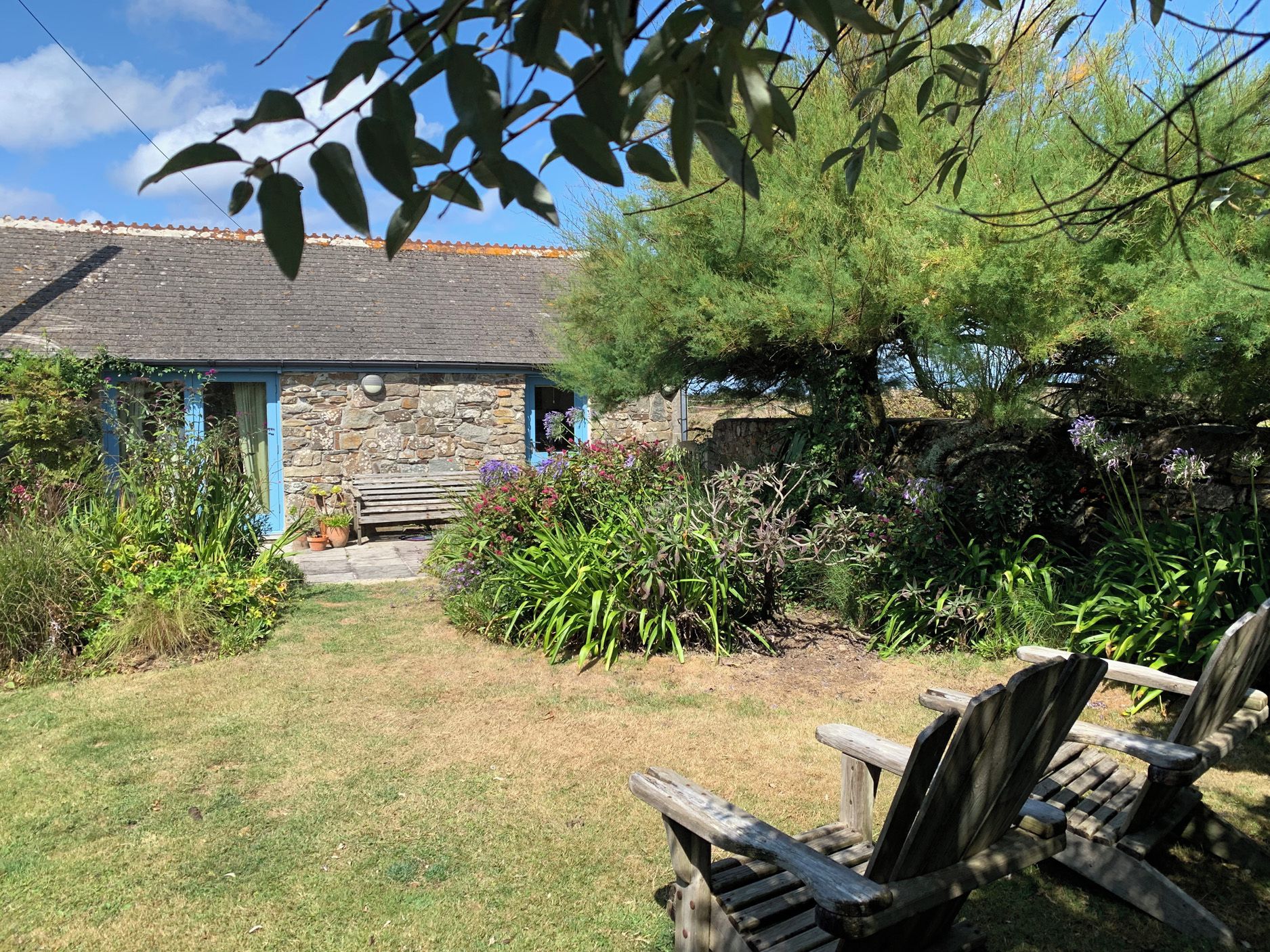 A stone building with a blue door, surrounded by a garden with various plants and a wooden bench.