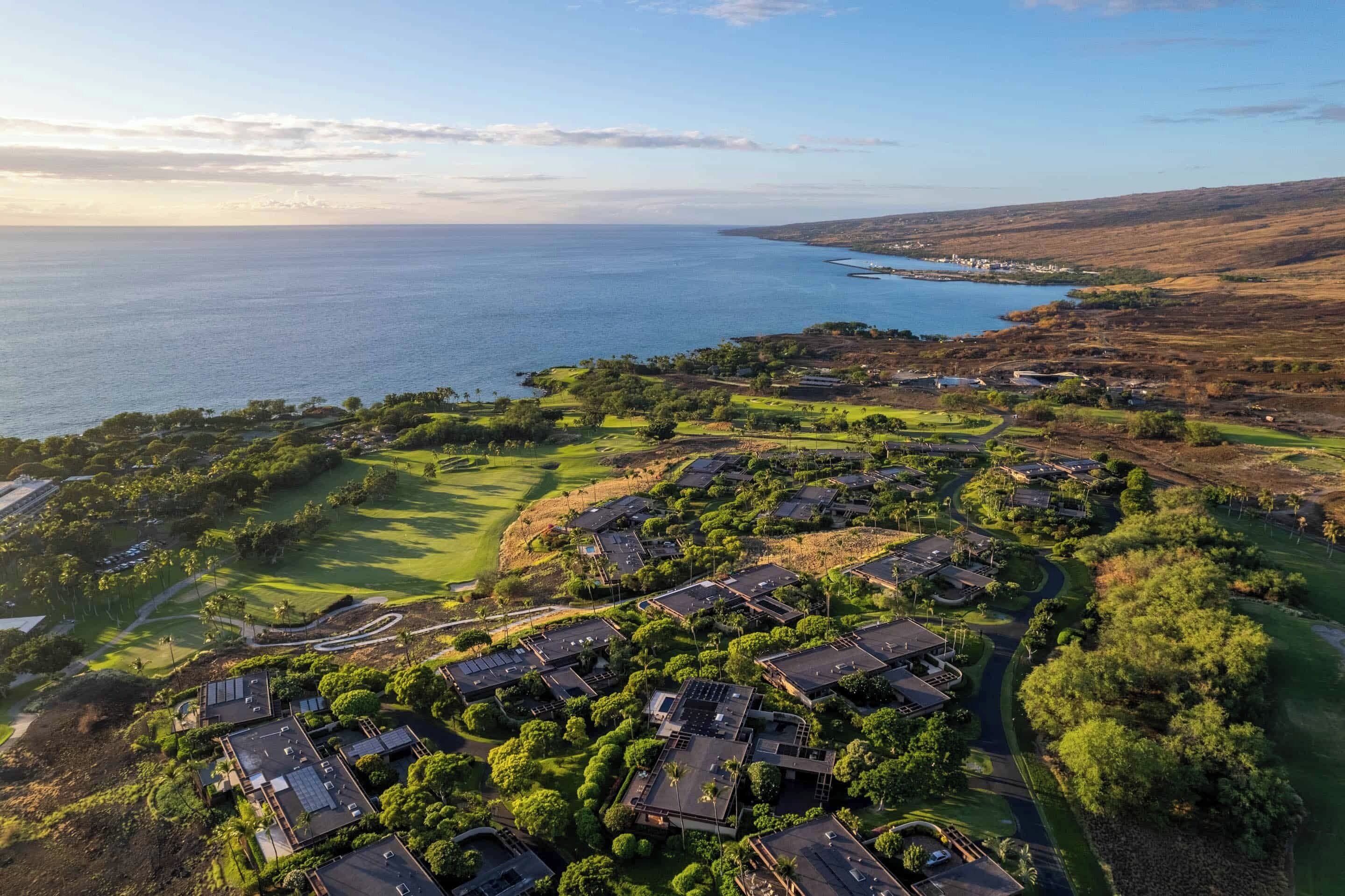 An aerial view of a coastal residential area with houses, a golf course, and a view of the ocean.