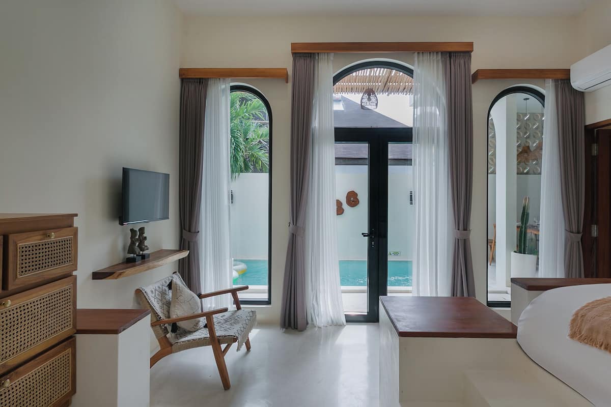 Une chambre avec un lit, une télévision, une commode en bois, une chaise et une vue sur une piscine, visible à travers une porte vitrée.
