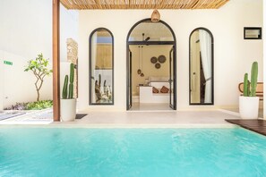 Zona de piscina con vista despejada a un baño que incluye bañera, ducha e inodoro. Hay cactus en macetas y un pequeño árbol cerca de la piscina.