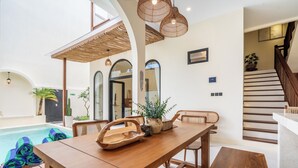 Amplio espacio interior con una mesa de comedor de madera, sillas y una piscina. Hay plantas en macetas y una escalera que conduce a otro nivel.