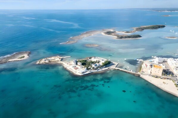 Vista aérea de una zona costera con aguas azules cristalinas, una pequeña isla con edificios y tierra firme con una estructura fortificada.