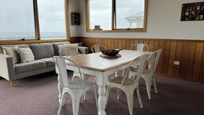 A dining area with a wooden table and white chairs, a grey sofa with cushions, and large windows with a view of the sea. - Drift Away with the waves and endless sea views (Greens Beach)