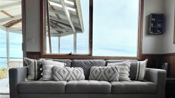 A grey sofa with patterned cushions in a room with a large window overlooking the sea. - Drift Away with the waves and endless sea views (Greens Beach)