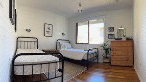 A small, tidy room with two metal beds, a wooden dresser, a large window, and a framed picture on the wall. - Drift Away with the waves and endless sea views (Greens Beach)