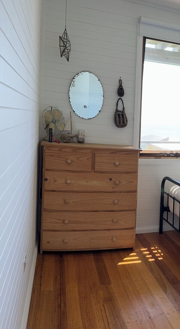 Une commode en bois avec des tiroirs, un miroir rond et un sac suspendu, le tout placé devant un mur blanc à panneaux.
