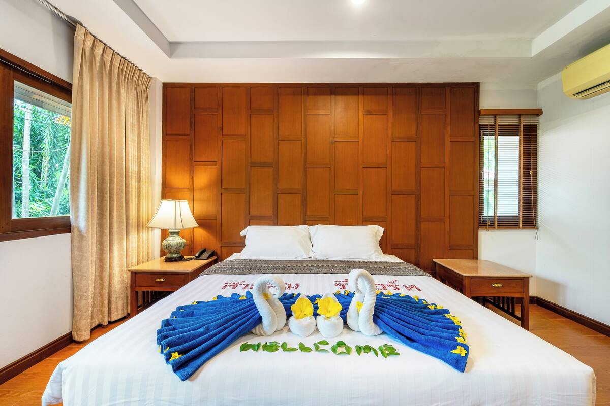 A hotel room with a neatly made bed, wooden headboard, a nightstand with a lamp, and a window with curtains.