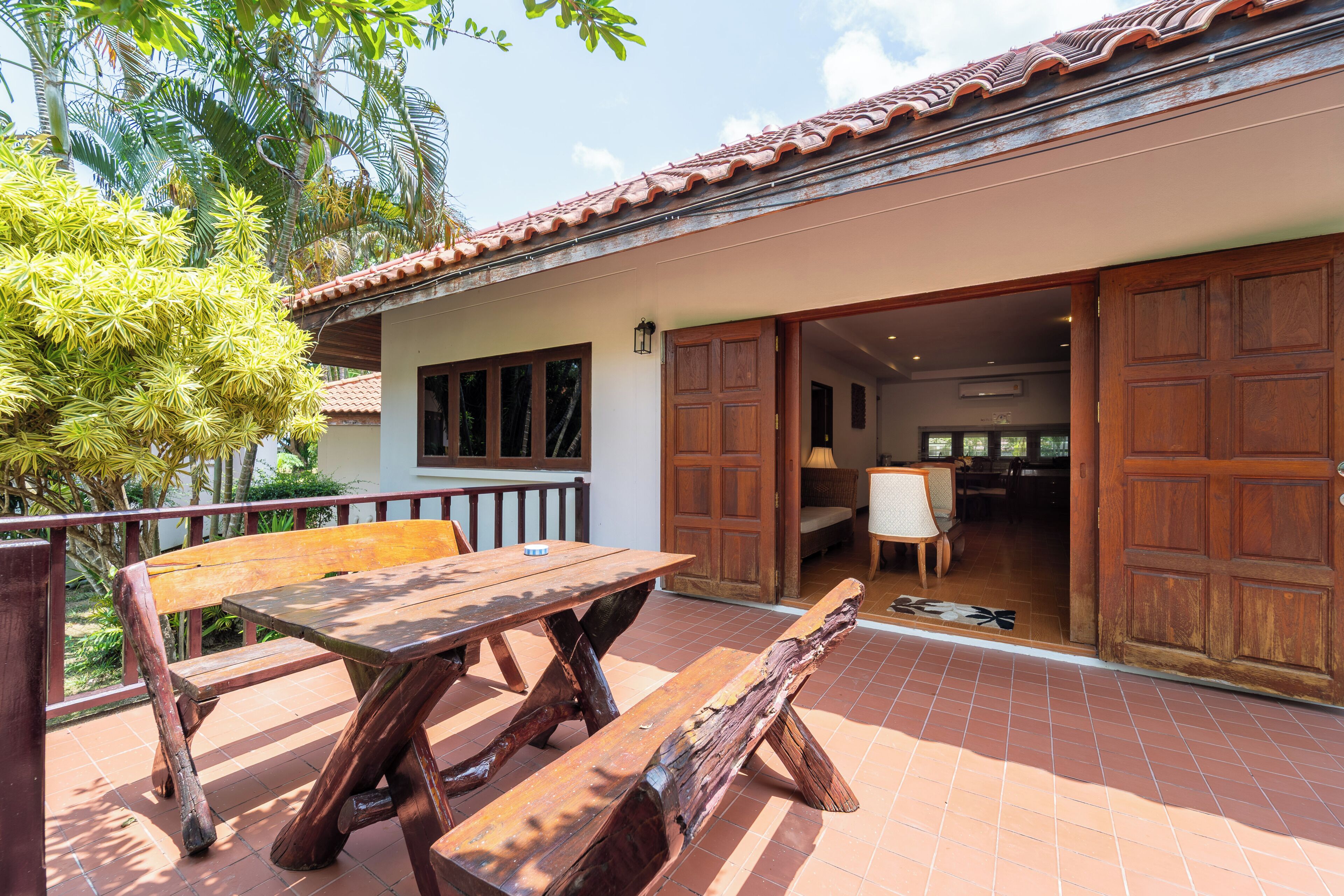 A patio with a wooden table and benches, a sliding door to a room, and a view of a garden with palm trees.