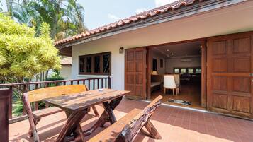 A patio with a wooden table and benches, a sliding door to a room, and a view of a garden with palm trees.