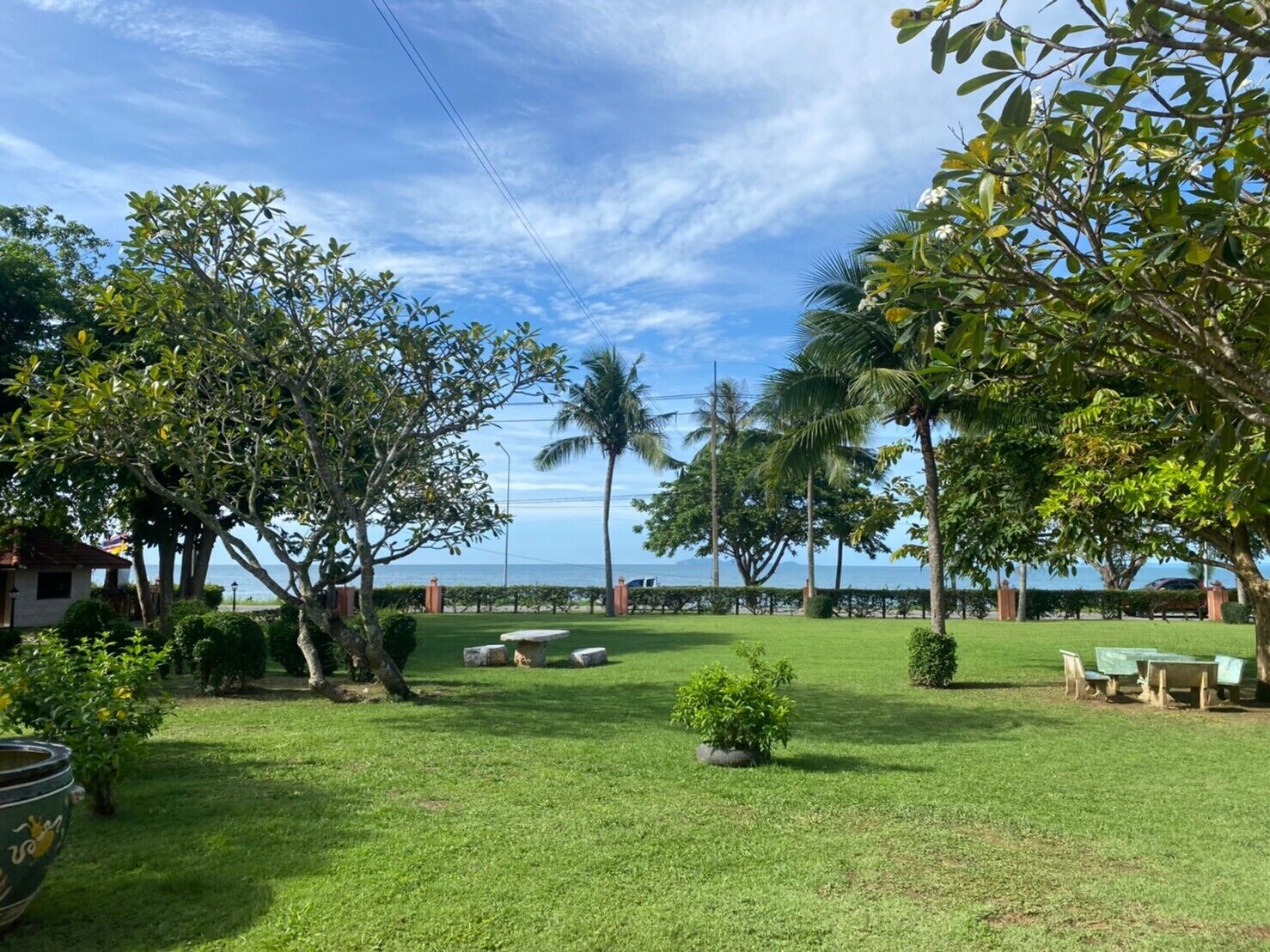 Um espaço ao ar livre bem cuidado, com gramado, várias árvores e vista para o mar. Há mesas de piquenique e um banco de pedra.