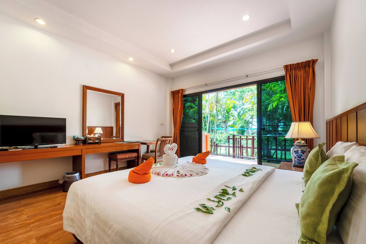 A hotel room with a bed, a desk with a television, a chair, a window with curtains, and a view of greenery outside.