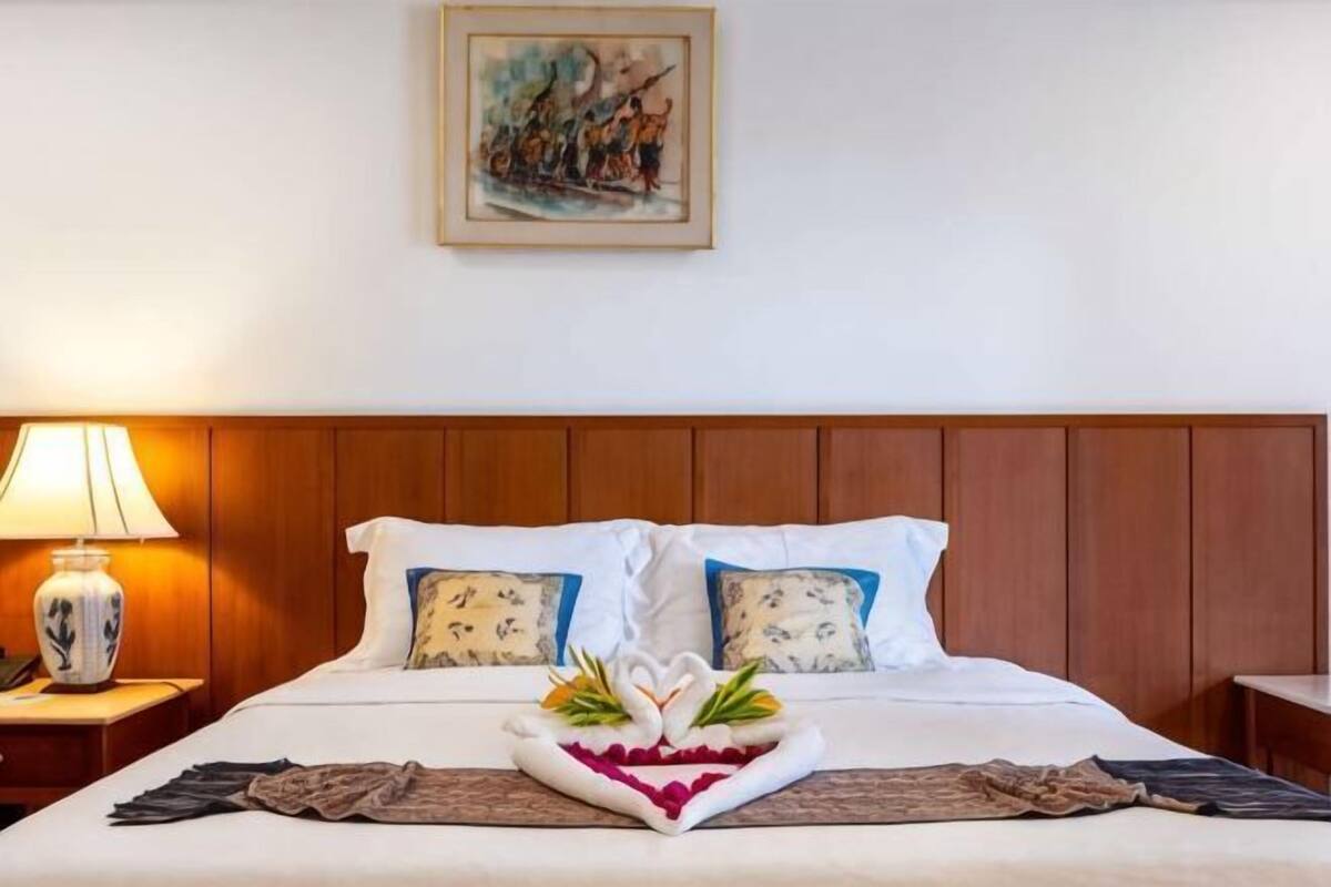 A hotel room with a wooden headboard, a bed with white linens, a decorative pillow, a bedside table with a lamp, and a painting on the wall.