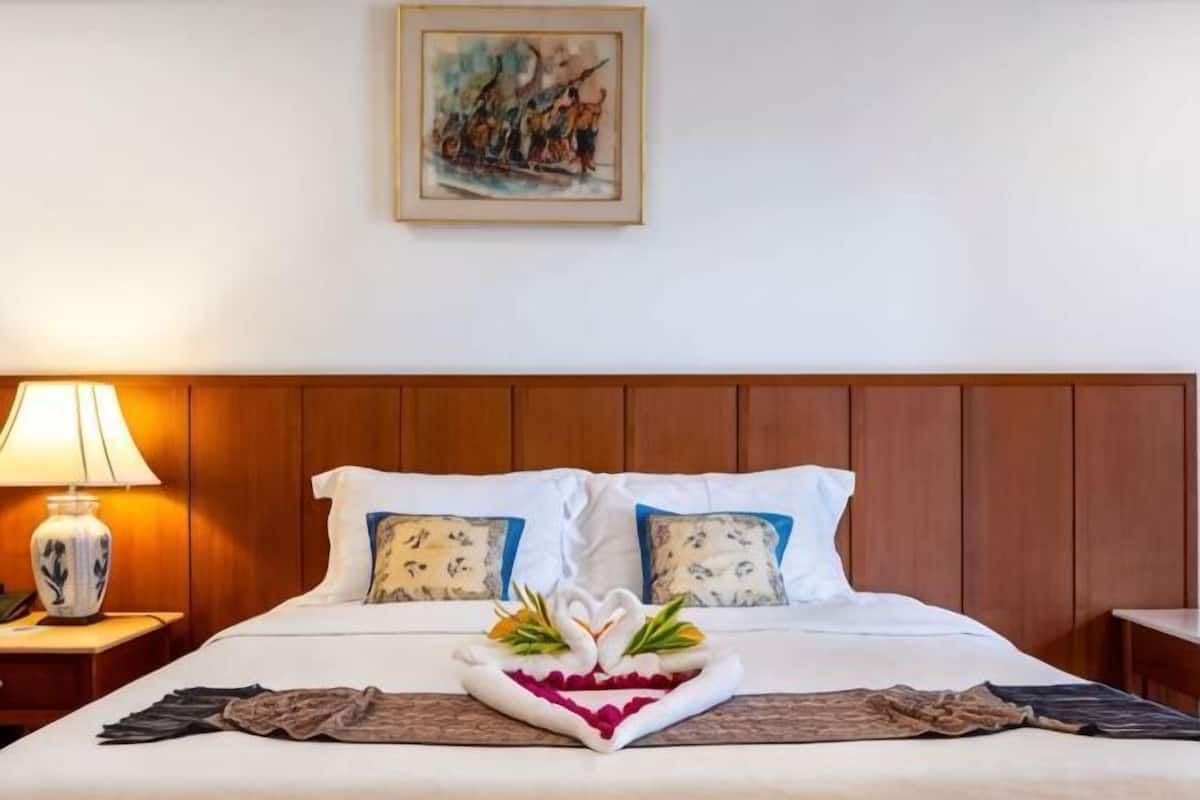 Ett hotellrum med en sänggavel i trä, en säng med vita sängkläder, en dekorativ kudde, ett nattduksbord med en lampa och en tavla på väggen.