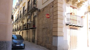 Una calle estrecha con un coche aparcado, edificios con balcones y un cartel rojo en la pared.