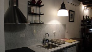 Una cocina con un lavamanos de acero inoxidable, encimera negra, salpicadero de mármol, estante negro con teteras rojas, una lámpara colgante y una pared blanca con un cuadro enmarcado.