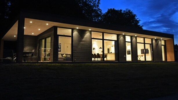 Une maison moderne dotée de grandes fenêtres et d’un toit plat, entourée d’arbres.