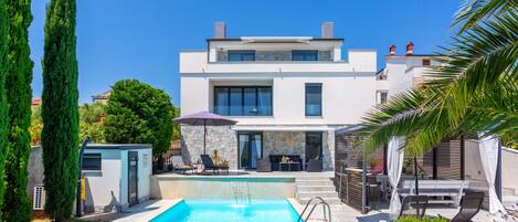 Une maison moderne à deux étages, dotée d’une piscine, d’un espace de détente extérieur et d’une façade en pierre.
