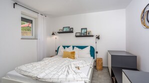 Un dormitorio con cama, ventana, mesita de noche y estante con fotografías.