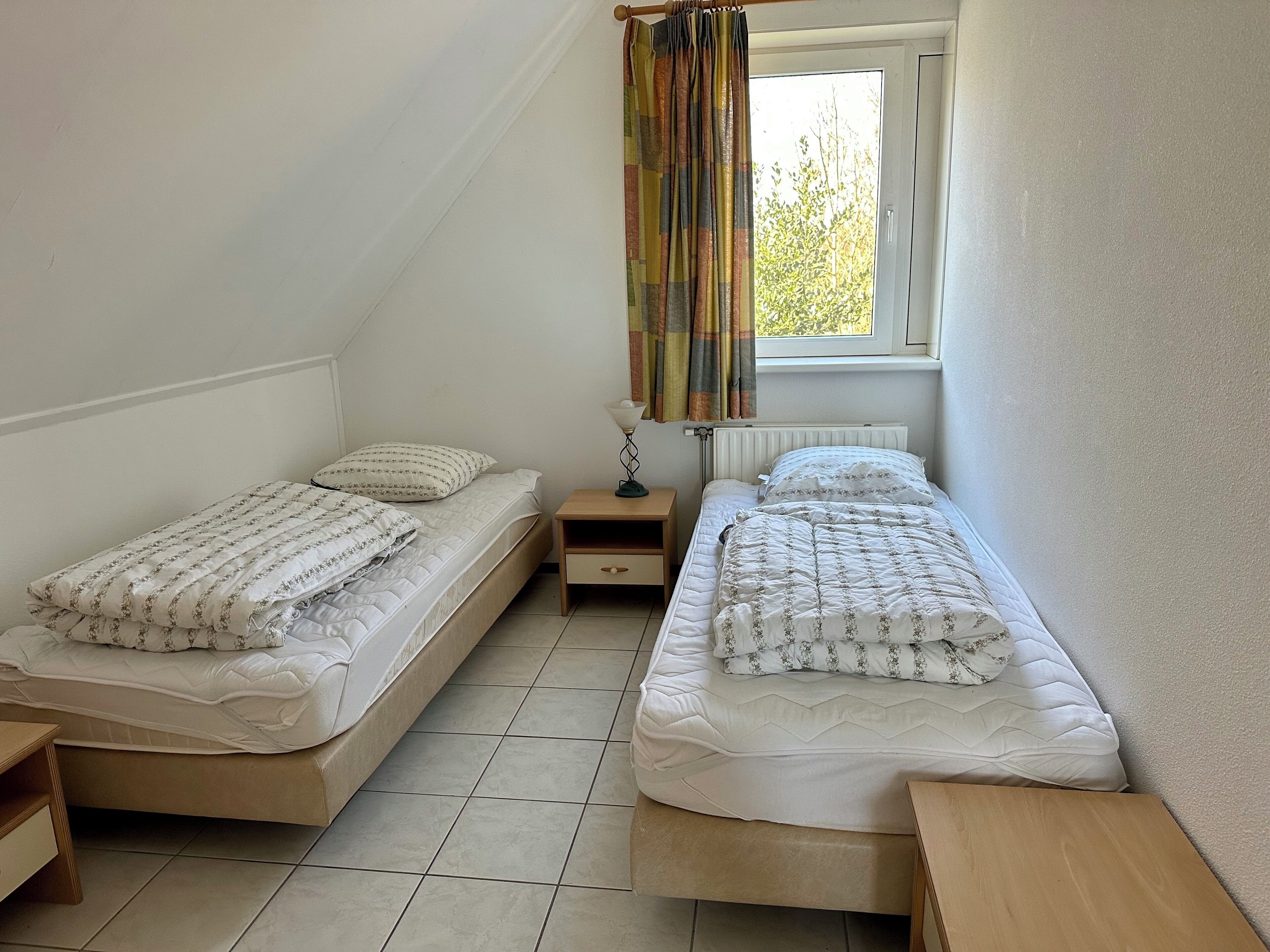 A small, clean room with two single beds, a nightstand, a lamp, and a window with curtains.