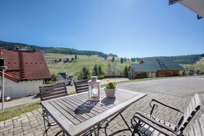 Une table en bois avec des bancs, une lanterne et une plante en pot, le tout installé dans un espace extérieur ensoleillé, avec vue sur un paysage vallonné.