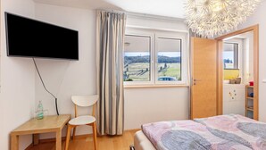 Une chambre à coucher avec un lit, un téléviseur fixé au mur, une fenêtre avec des rideaux, un bureau en bois avec une chaise, et une vue sur un paysage.