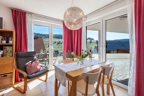 A modern room with a dining table, chairs, a sofa, and a large window offering a view of the countryside.