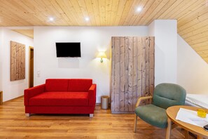Una habitación con un sofá rojo, una silla verde, una mesa de madera, un televisor montado en la pared y un armario de madera.
