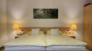 Un lit double avec une tête de lit en bois, deux lampes de chevet, une table de chevet avec une lampe et un tableau représentant une forêt, encadré, accroché au mur.