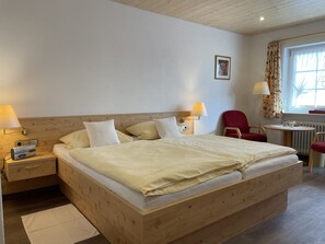 Een hotelkamer met een houten bed, twee nachtkastjes met lampen, een rode stoel, een klein tafeltje met een lamp, en een raam met gordijnen.