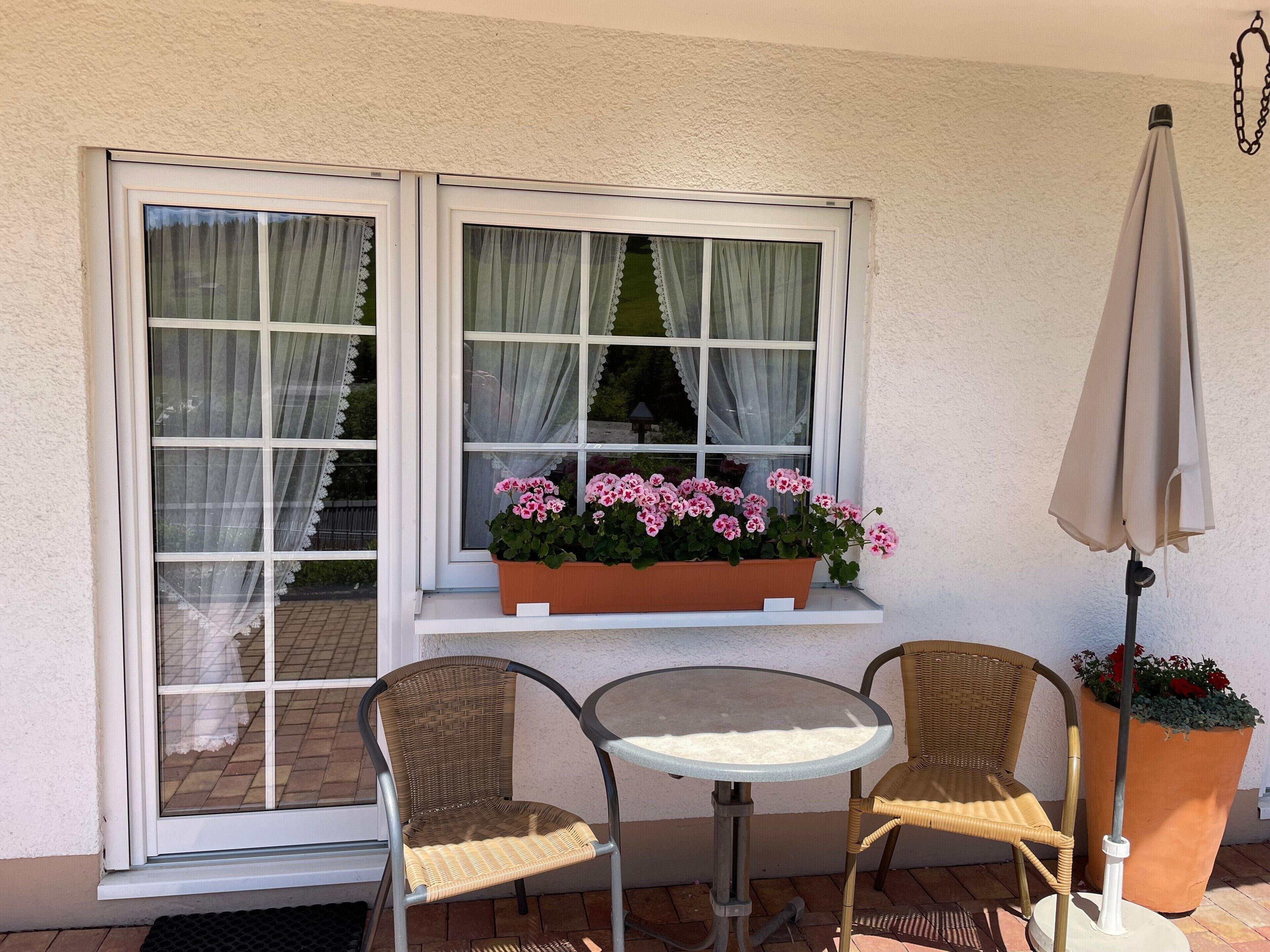 Un patio con una mesa y dos sillas, una ventana con cortinas, una planta en maceta con flores rosadas y un paraguas de pie.