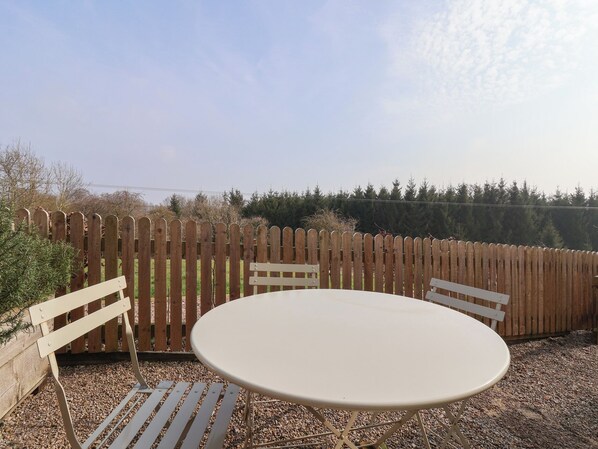 Une table ronde avec deux chaises, située dans un espace jardin agrémenté d’une clôture en bois et bordé d’arbres.