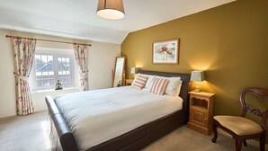Een slaapkamer met een groot bed, een nachtkastje met een lamp, een stoel, een raam met gordijnen en een schilderij aan de muur.