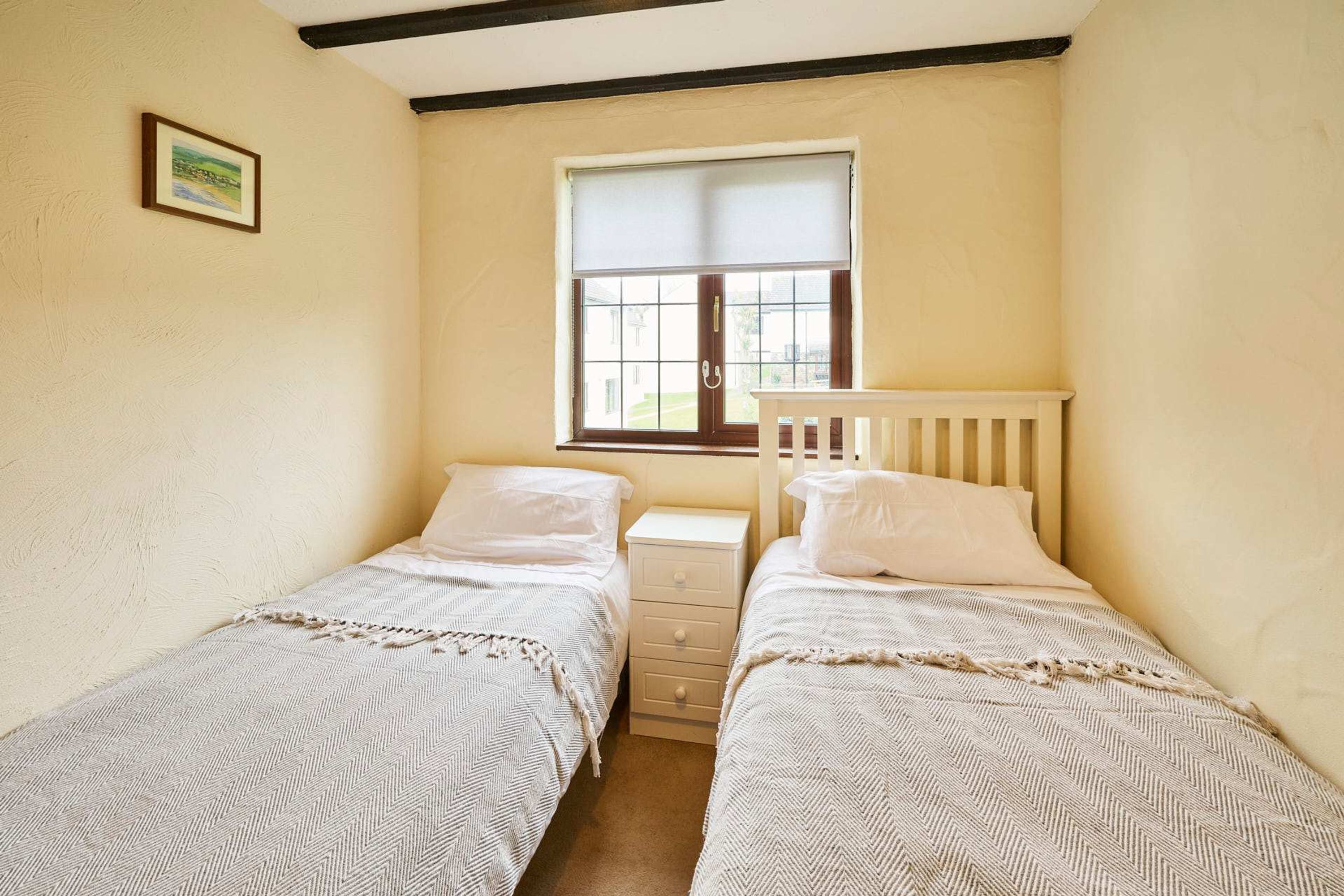 A small, cozy bedroom with two single beds, a nightstand, a window with a view, and a framed picture on the wall.