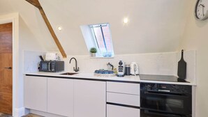 Une cuisine moderne avec des meubles blancs, un four encastré, un four à micro-ondes et une machine à café. Il y a une fenêtre au-dessus de l’évier et une horloge au mur.