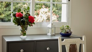 Een raam met uitzicht op groen, een vaas met rode bloemen, een lantaarn en een blauwe kom met planten op een grijze dressoir.