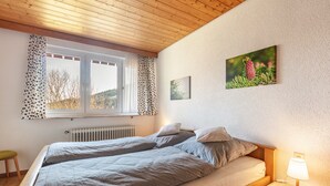 A bedroom with two beds, a window with curtains, a radiator, a bedside table with a lamp, and a framed picture on the wall. - Apartment Eddy, 45 sqm with Mountain View, Meadow and Wi-Fi (Todtnau)