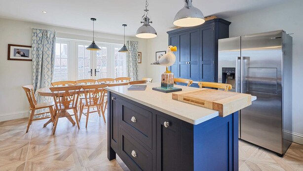 A modern kitchen with a central island, dark cabinetry, stainless steel appliances, and a dining area with wooden chairs and a table. - Host Stay Kittiwake Cottage (Bridlington)
