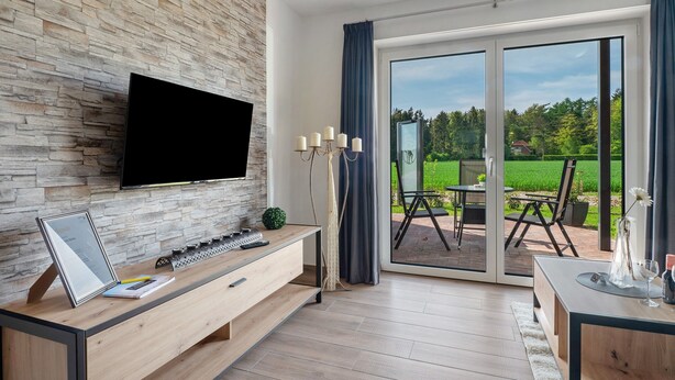 A modern room with a stone wall, a flat-screen TV, a wooden desk, and a sliding glass door leading to an outdoor patio. - Holiday Apartment 'Anna 1 Eg' with Private Terrace and Garden (Suhle)