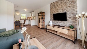A modern living room with a flat-screen TV, wooden console, and a dining area with a kitchen in the background. - Holiday Apartment 'Anna 1 Eg' with Private Terrace and Garden (Suhle)